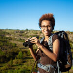 Young beautiful african girl with backpack smiling, looking at camera, holding binoculars, canyon background. Copy space.